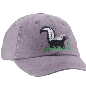 May include: A purple baseball cap with an embroidered skunk design. The skunk is black and white with a white stripe down its back. It is standing on a green patch of grass with a pink flower.