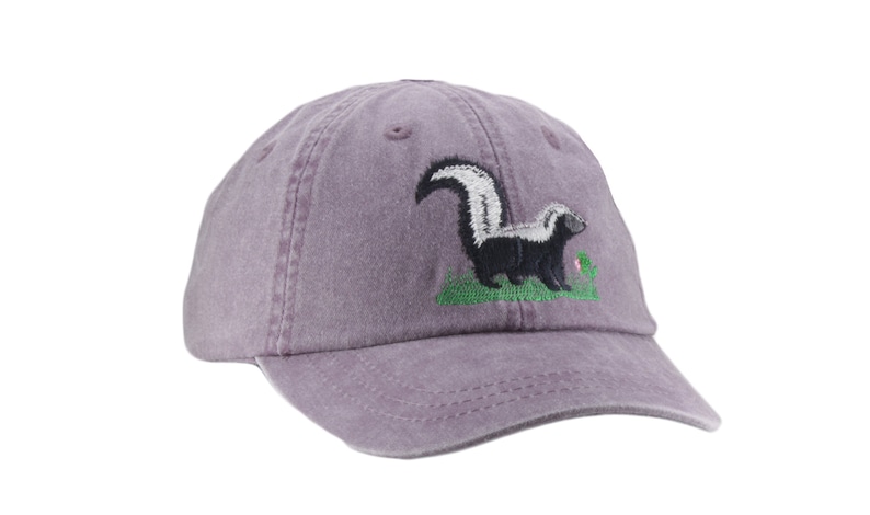 May include: A purple baseball cap with an embroidered skunk design. The skunk is black and white with a white stripe down its back. It is standing on a green patch of grass with a pink flower.