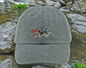 May include: A dark green baseball cap with an embroidered fishing fly on the front. The fly is black, white, and red with a gold hook.