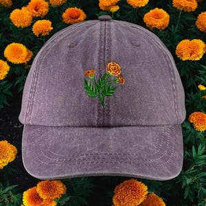 May include: A purple denim baseball cap with an embroidered design of orange marigolds.