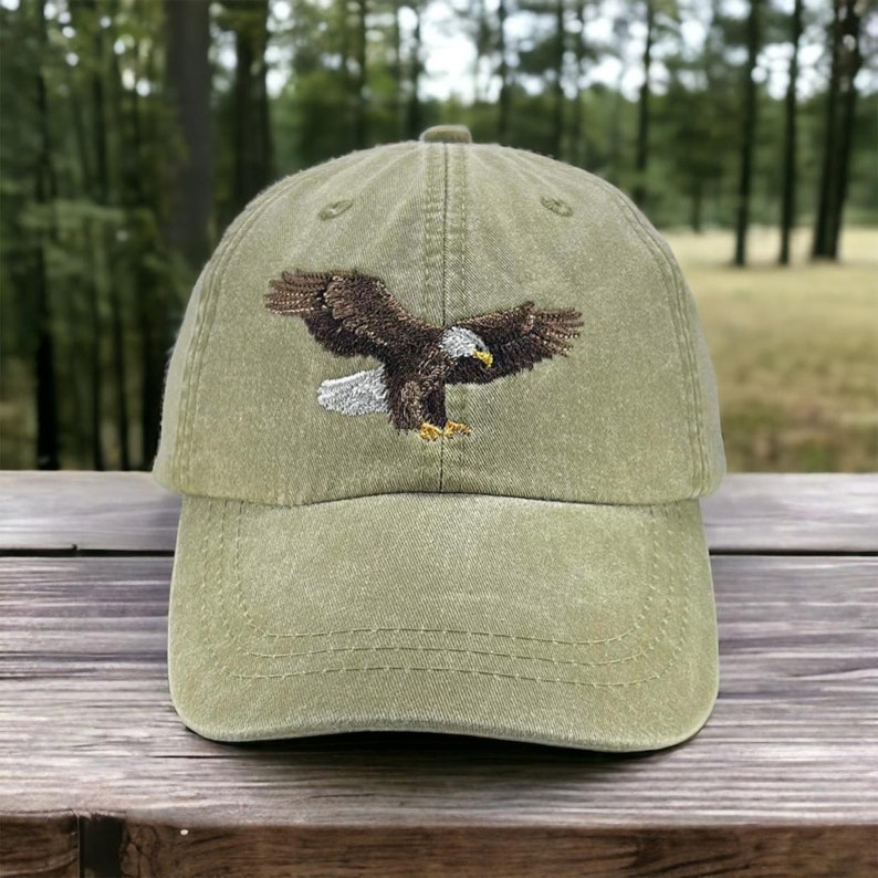 May include: Olive green baseball cap with an embroidered eagle design. The eagle is depicted with brown and white feathers, and a yellow beak. The cap has a curved brim and an adjustable strap.