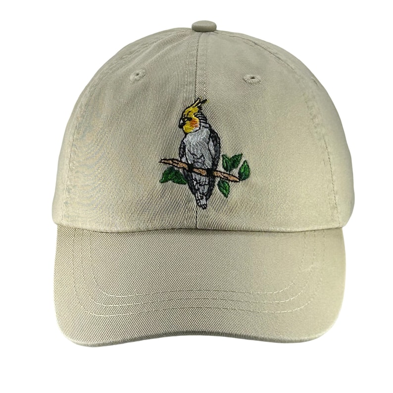 May include: A beige baseball cap with an embroidered image of a cockatiel perched on a branch.