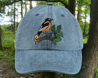 May include: A blue denim baseball cap with an embroidered image of an orange and black bird perched on a branch with green leaves.