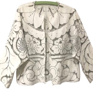 May include: A white open-front jacket featuring a dark grey floral and botanical line-art pattern throughout. The garment has a round neckline, wide three-quarter length sleeves, and a straight hemline, showcasing a detailed illustrative design.