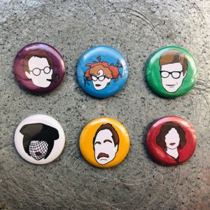 May include: Six round button pins featuring colorful illustrations of different people. The pins are approximately 1 inch in diameter.