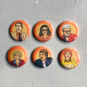 May include: Six round orange and white button pins featuring portraits of six different people. The pins are arranged in two rows of three.