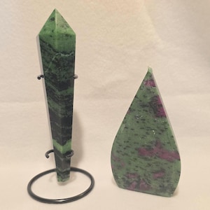 May include: A tall, polished green and black stone obelisk held by a black metal wire stand, positioned next to a flat, teardrop-shaped stone carving featuring a mottled green surface with distinct patches of deep magenta and black inclusions.