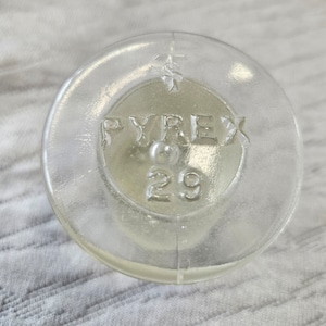 Puede incluir: Un tapón de botella de vidrio transparente con la palabra "PYREX" y el número "29" en relieve.