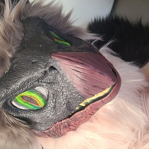 以下が含まれることがあります： 灰色とピンクの毛皮で覆われた動物の頭で、黒い鼻と緑色の目があります。頭には、赤色の口をした黒と灰色の恐竜のようなマスクが付いています。