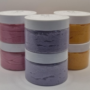 May include: Six clear plastic jars with white screw-top lids arranged in three stacks of two. The jars contain textured body scrub in pink, purple, and orange colors. Each lid features a circular white label with black text and a logo.