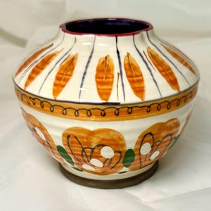 May include: A white ceramic vase with a rounded shape and a decorative design. The vase features orange, green, and white painted details, including stylized leaves and flowers.