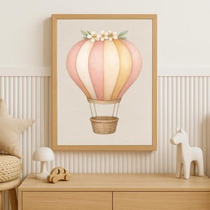 Könnte beinhalten: Aquarell-Illustration eines Heißluftballons in einem Holzrahmen. Der Ballon hat rosa, gelbe und weiße Streifen, einen Weidenkorb und Blumendetails. Ein charmantes Stück für die Kinderzimmerdekoration.