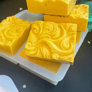 May include: Four yellow soap bars with a swirl design in a clear silicone mold. The soap bars are stacked on top of each other.
