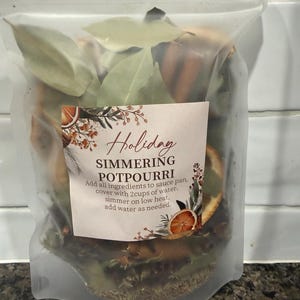 May include: A clear plastic pouch containing dried holiday simmering potpourri, including bay leaves and orange slices. The label reads: Holiday SIMMERING POTPOURRI. Add all ingredients to sauce pan, cover with 2cups of water, simmer on low heat, add water as needed.