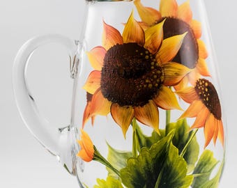 May include: A clear glass pitcher with a painted design of sunflowers and green leaves. The pitcher has a handle and a wide mouth.