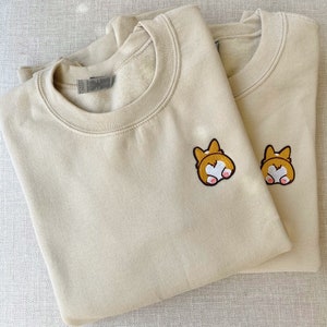 Könnte beinhalten: Zwei beigefarbene Sweatshirts mit gestickten Bildern eines Corgi-Hinterteils. Die Corgis sind vom Betrachter abgewandt und haben einen herzförmigen Schwanz.