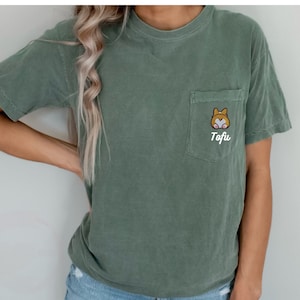 Könnte beinhalten: Ein grünes T-Shirt mit einer Tasche, auf der ein Cartoon-Corgi mit dem Namen "Tofu" gestickt ist.