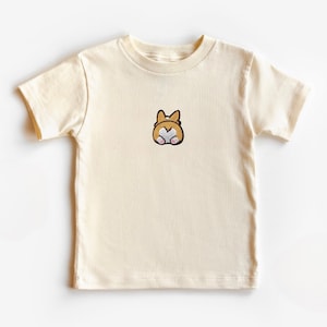Könnte beinhalten: Cremefarbenes T-Shirt mit einem kleinen, gestickten Design eines Corgi-Hinterns. Das Design umfasst braune, weiße und rosa Details. Das Shirt besteht aus weichem Material und hat kurze Ärmel.
