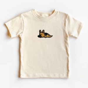 May include: A cream-colored short-sleeved t-shirt with an embroidered image of a brown and black German Shepherd dog sleeping.