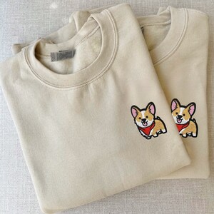 Könnte beinhalten: Zwei beigefarbene Sweatshirts mit Rundhalsausschnitt und gestickten Bildern eines Corgis mit rotem Halstuch.