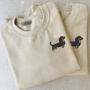 Pode incluir: Duas sweatshirts bege com cães dachshund bordados. Os cães são pretos com marcas marrons e estão virados para a direita.