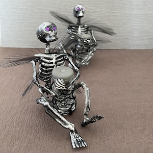 May include: Two silver skeleton figurines with purple eyes. One is playing a drum and the other is holding a drum stick.