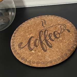 May include: A round cork coaster with a decorative border and the word "coffee" written in cursive with a steam design above it.
