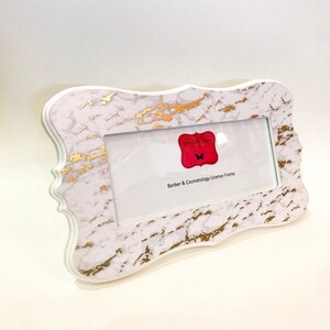 May include: White and gold marble-patterned license frame with a decorative edge. The frame has a rectangular opening for a license and a red label with the text "Familiar Faces" and "Barber & Cosmetology License Frame".
