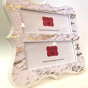 May include: A double-opening frame with a white and gold marble design. The frame has a decorative shape and includes the text "Barber & Cosmetology License Frame". The frame is designed to hold two photos or documents.