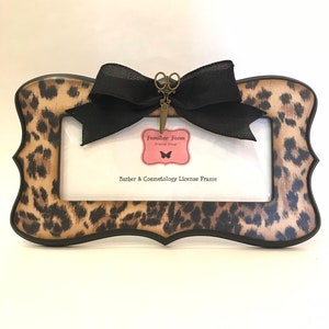 May include: A rectangular license frame with a leopard print design and a black border. A black bow and a scissors charm are attached to the frame. The frame has the text "Barber & Cosmetology License Frame". A pink tag with the text "Familiar Faces Frame Shop" is also attached.