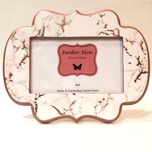 May include: A white and rose gold marble-effect license frame with a distinctive scalloped edge. The frame has a rose gold border and holds a 20 x 13 cm license. The frame includes the text "Familiar Faces Frame Shop" and "Barber & Cosmetology License Frame."