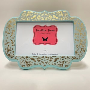 May include: A pale blue picture frame with gold floral detailing. The frame is shaped decoratively and holds a 20.3 x 12.7 cm photo. The frame has the text "Familiar Faces Frame Shop" and "Barber & Cosmetology License Frame".