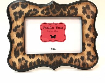 Puede incluir: Un marco de fotos rectangular con un diseño de estampado de leopardo. El marco tiene un borde negro y una forma decorativa. El marco tiene una etiqueta roja con el texto "Familiar Faces Frame Shop" y una mariposa. El marco mide 10 x 15 cm.