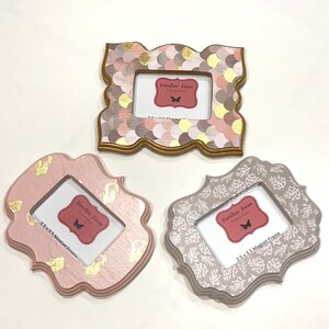 Op de afbeelding: Drie decoratieve magneetframes in verschillende ontwerpen. Eén frame heeft een roze, gouden en bruin geschubd patroon. Een andere is lichtroze met gouden accenten, en de derde is grijs met een wit koraalontwerp. Elk frame heeft een opening van 6,35 x 8,89 cm.