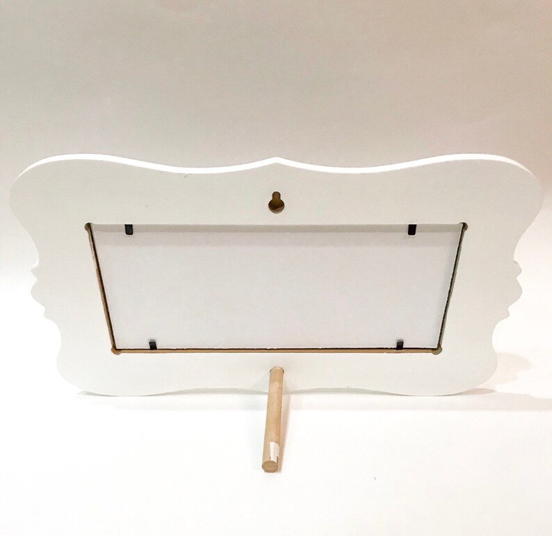 May include: A white, ornate picture frame with a decorative silhouette design on the sides. The frame has a rectangular opening and a wooden stand. A small metal hook is attached to the back for hanging.