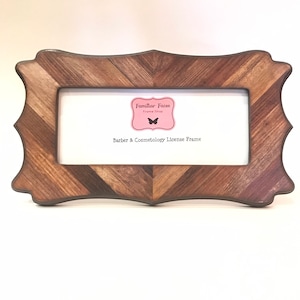 May include: A wooden frame with a chevron pattern in shades of brown and grey. The frame has a decorative, scalloped edge and a white interior. The text "Barber & Cosmetology License Frame" is visible.