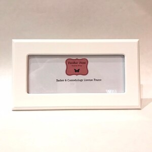 May include: A white rectangular frame with a rectangular opening. The frame has the text "Barber & Cosmetology License Frame" below a logo that says "Familiar Faces Frame Shop".