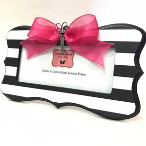 May include: A black and white striped frame with a pink bow and a scissors charm. The frame has a decorative shape and holds a white space for a license. Text on the frame reads "Barber & Cosmetology License Frame".