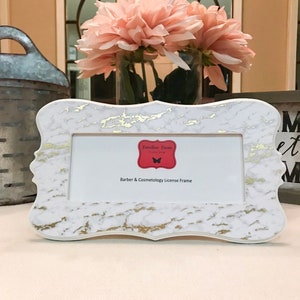 May include: A white and gold marble-patterned license frame with a decorative shape. The frame has a red label with the text "Familiar Faces" and "Barber & Cosmetology License Frame". A vase of pink flowers is in the background.