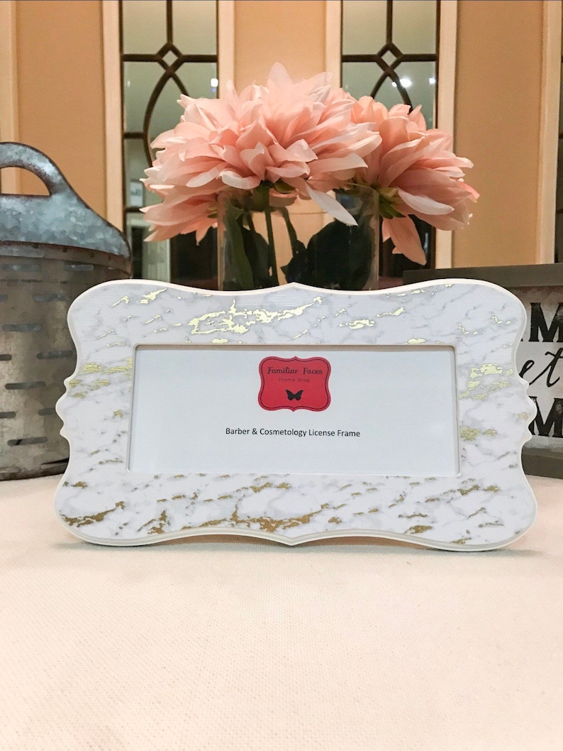 May include: A white and gold marble-patterned license frame with a decorative shape. The frame has a red label with the text "Familiar Faces" and "Barber & Cosmetology License Frame". A vase of pink flowers is in the background.