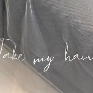 Pode incluir: Um close-up de um véu cinza com as palavras "Take my hand" bordadas em escrita branca. O véu tem uma textura macia e transparente e é ligeiramente translúcido, com o texto visível na superfície.