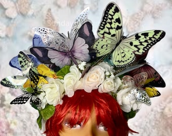 May include: A white flower crown with butterflies and greenery. The crown is made of artificial flowers and butterflies. The butterflies are in various colors, including purple, green, and silver.