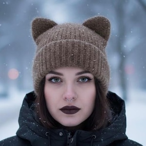 Könnte beinhalten: Eine braune Strickmütze mit Katzenohren. Die Person trägt eine schwarze Jacke. Der Hintergrund ist verschwommen mit einem Schneeeffekt.