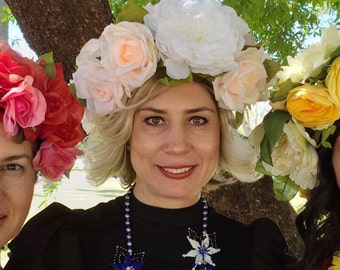 May include: A woman wearing a floral crown of white and peach-colored roses and a blue and white beaded necklace. The woman is smiling, with a tree trunk in the background. Other women wearing floral crowns are visible.