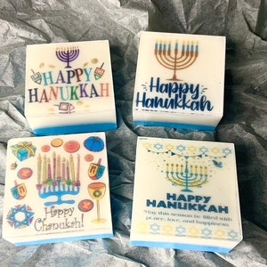 May include: Four white square soaps with blue trim, each featuring a different Hanukkah design. The top left soap has a menorah with candles and the text "HAPPY HANUKKAH". The top right soap has a menorah with candles and the text "Happy Hanukkah". The bottom left soap has a menorah with candles, dreidels, and the text "Happy Chanukah!" The bottom right soap has a menorah with candles, stars, and the text "HAPPY HANUKKAH" with the message "May this season be filled with peace, love, and happiness."