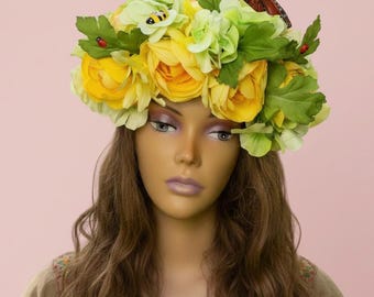 May include: A floral crown featuring yellow roses, green hydrangeas, and fabric leaves. A monarch butterfly and small insect accents the crown. The crown is worn by a mannequin with long brown hair, against a pink background.