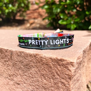May include: Black fabric wristband with the words "PRETTY LIGHTS" in white. The band has a colorful design with musical notes, stars, and a heartbeat graphic. The background is a blurred green and the wristband is resting on a brown stone.