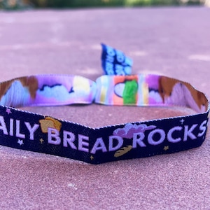May include: A black and blue woven wristband with the text "DAILY BREAD ROCKS" in white lettering. The wristband has a colourful design with clouds, stars, and bread.