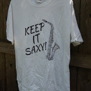 Puede incluir: Camiseta blanca con un gráfico negro de un saxofón y el texto "Keep It SaxY!"