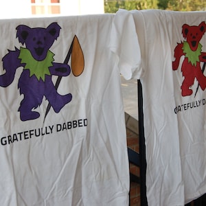May include: Two white t-shirts with a graphic print of a cartoon bear holding a lightning bolt. The text "Gratefully Dabbed" is printed below the bear on each shirt.